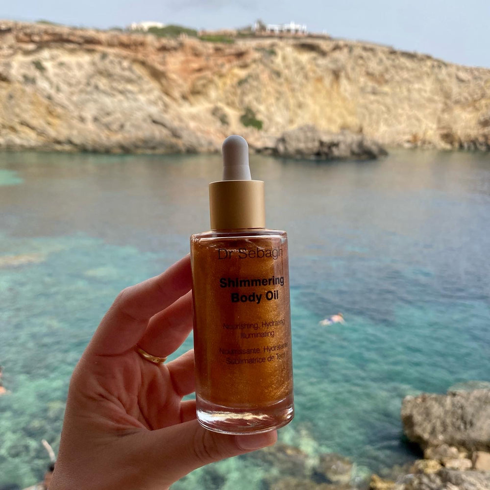 Hand holding a bottle of 'Shimmering Body OIl with a scenic background of water and cliffs.