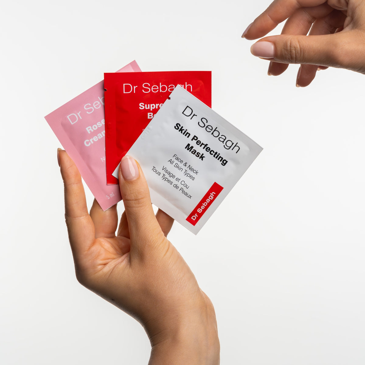 Three Dr Sebagh Mature Skin Discovery Kit skincare packet sachets in pink, red, and white are held between two hands against a clean white background.