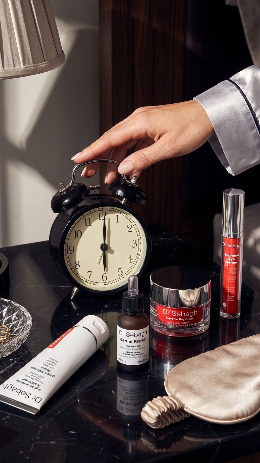 Dr Sebagh skincare products arranged on a dark bedside table with an alarm clock, silk eye mask and a hand reaching across in morning light