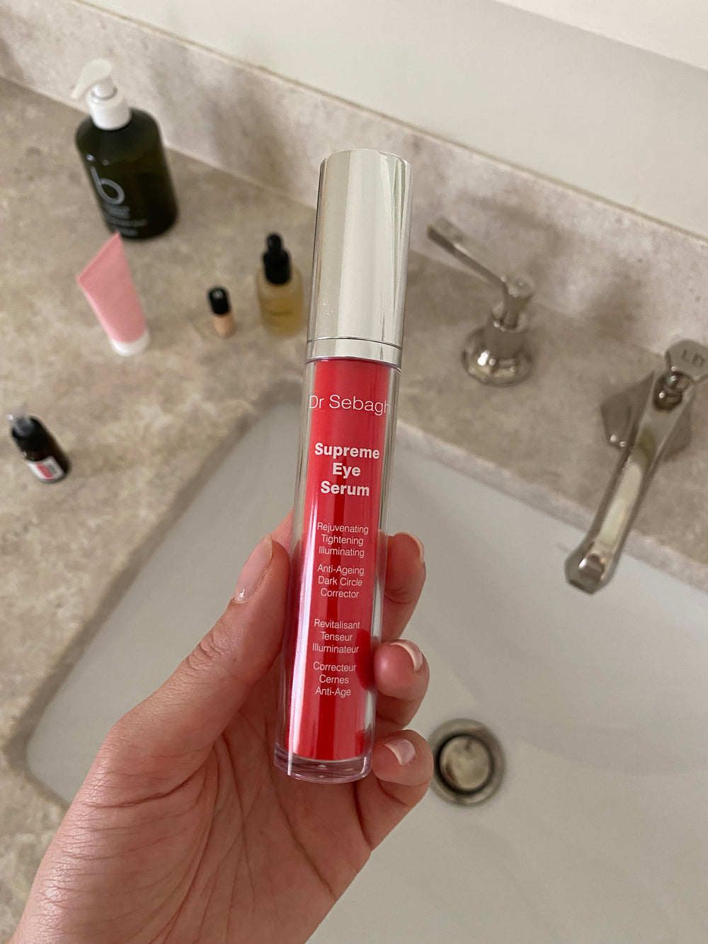 Hand holding a red Dr Sebagh Supreme Eye Serum bottle with visible text, against a bathroom counter background.